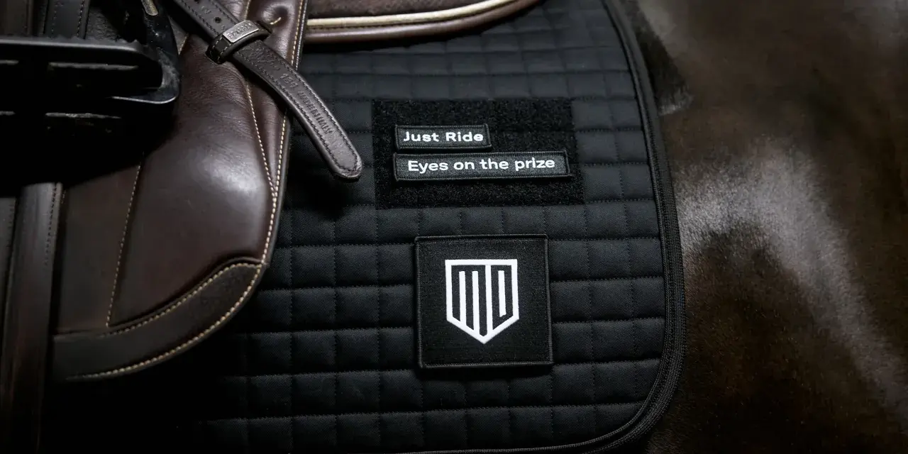 Schwarze Schabracke mit Aufnähern "Just Ride" und "Eyes on the prize". Die Schabracke ist Teil der Reitausrüstung und unterstreicht die Bedeutung von Fokus und Zielstrebigkeit.