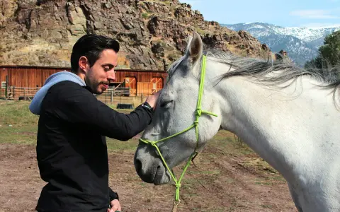 Mann streichelt Pferd, zeigt Vertrauen und Harmonie im **natural horsemanship**. Im Hintergrund eine rustikale Scheune und Berge.