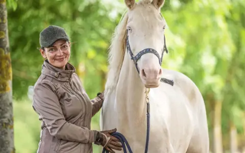 Frau mit Kappe und brauner Jacke streichelt ein helles Pferd. Sie hält eine blaue Leine. Faszientraining Pferd für mehr Wohlbefinden.