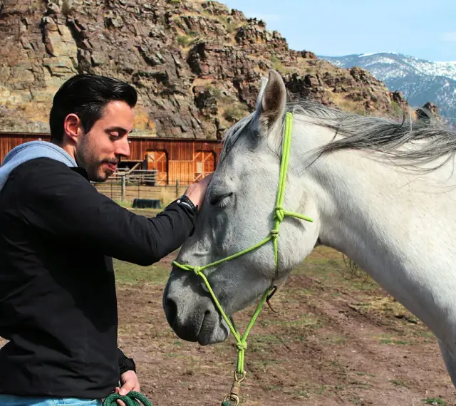 Mann streichelt Pferd, zeigt Vertrauen und Harmonie im **natural horsemanship**. Im Hintergrund eine rustikale Scheune und Berge.