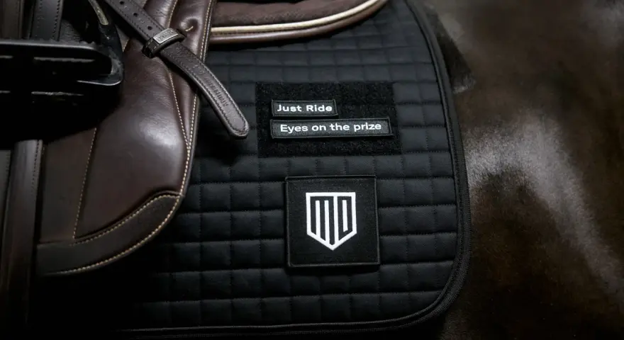 Schwarze Schabracke mit Aufnähern "Just Ride" und "Eyes on the prize". Die Schabracke ist Teil der Reitausrüstung und unterstreicht die Bedeutung von Fokus und Zielstrebigkeit.