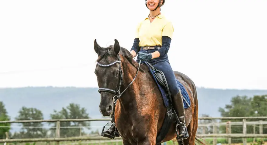 Eine Reiterin in gelbem Shirt und Reitstiefeln genießt das Aussitzen beim Reiten auf einem braunen Pferd.