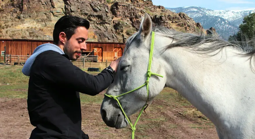 Mann streichelt Pferd, zeigt Vertrauen und Harmonie im **natural horsemanship**. Im Hintergrund eine rustikale Scheune und Berge.