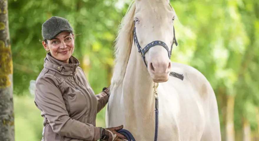 Frau mit Kappe und brauner Jacke streichelt ein helles Pferd. Sie hält eine blaue Leine. Faszientraining Pferd für mehr Wohlbefinden.