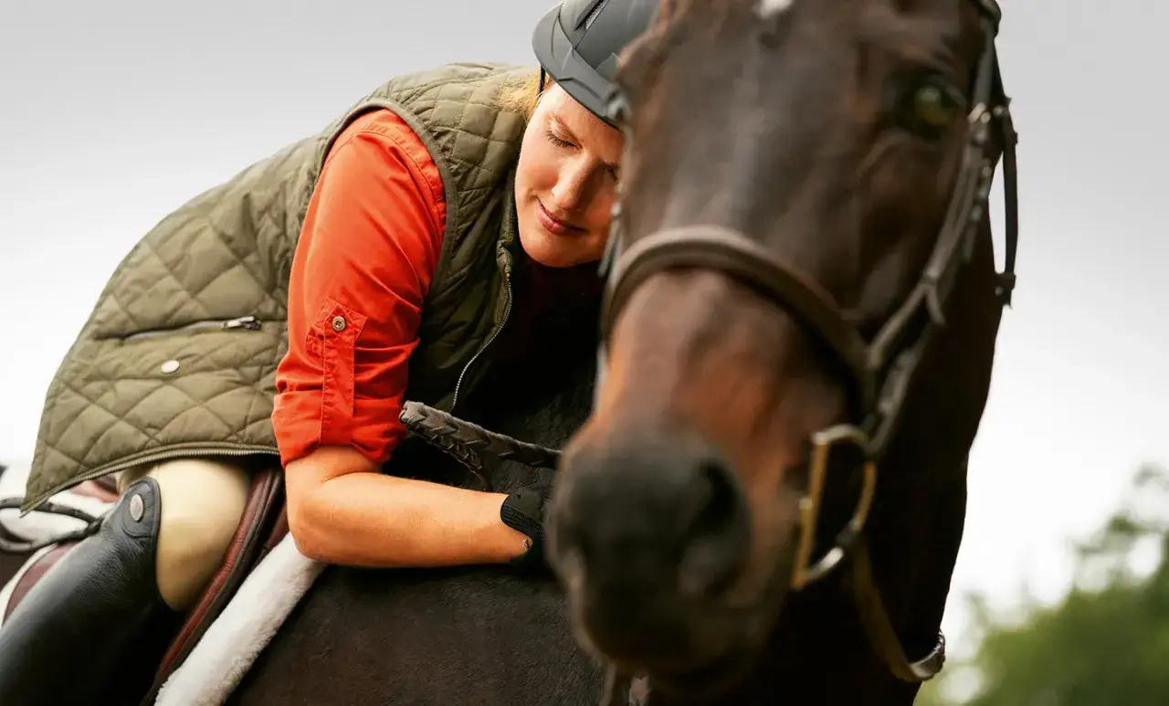 Eine Reiterin mit Helm und Weste lehnt sich vertrauensvoll an ihr Pferd. Die Szene zeigt die tiefe Verbindung beim Horsemanship Training.