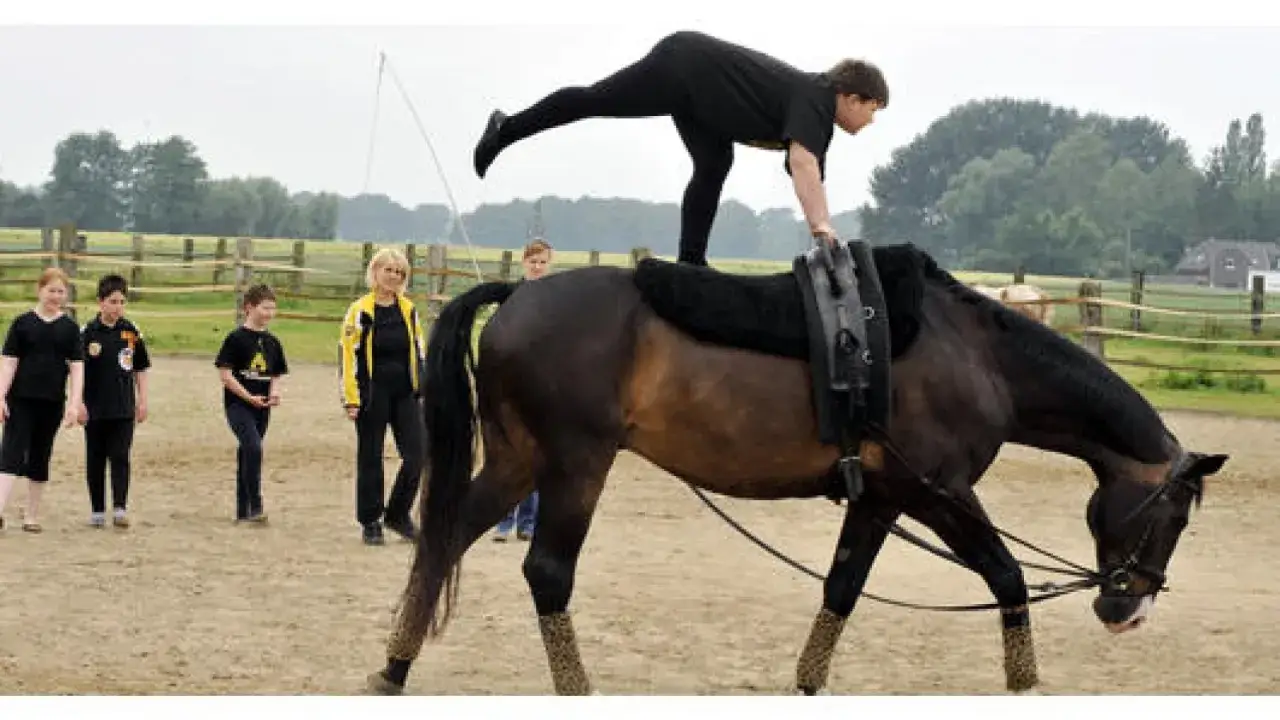 Akrobatin balanciert auf einem Pferd beim Voltigieren. Kinder und eine Trainerin beobachten gespannt.