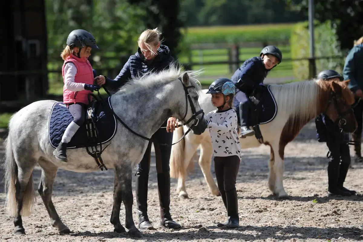 Kinder reiten auf Ponys, eine Frau hilft einem Mädchen auf dem grauen Pony.
