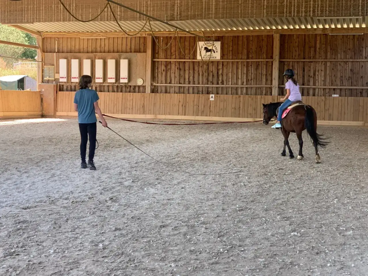 Ein Mädchen reitet ein Pferd in einer Reithalle, während eine andere Person sie an der Longe führt. Ob das Reiten schwer ist, zeigt sich im Training.