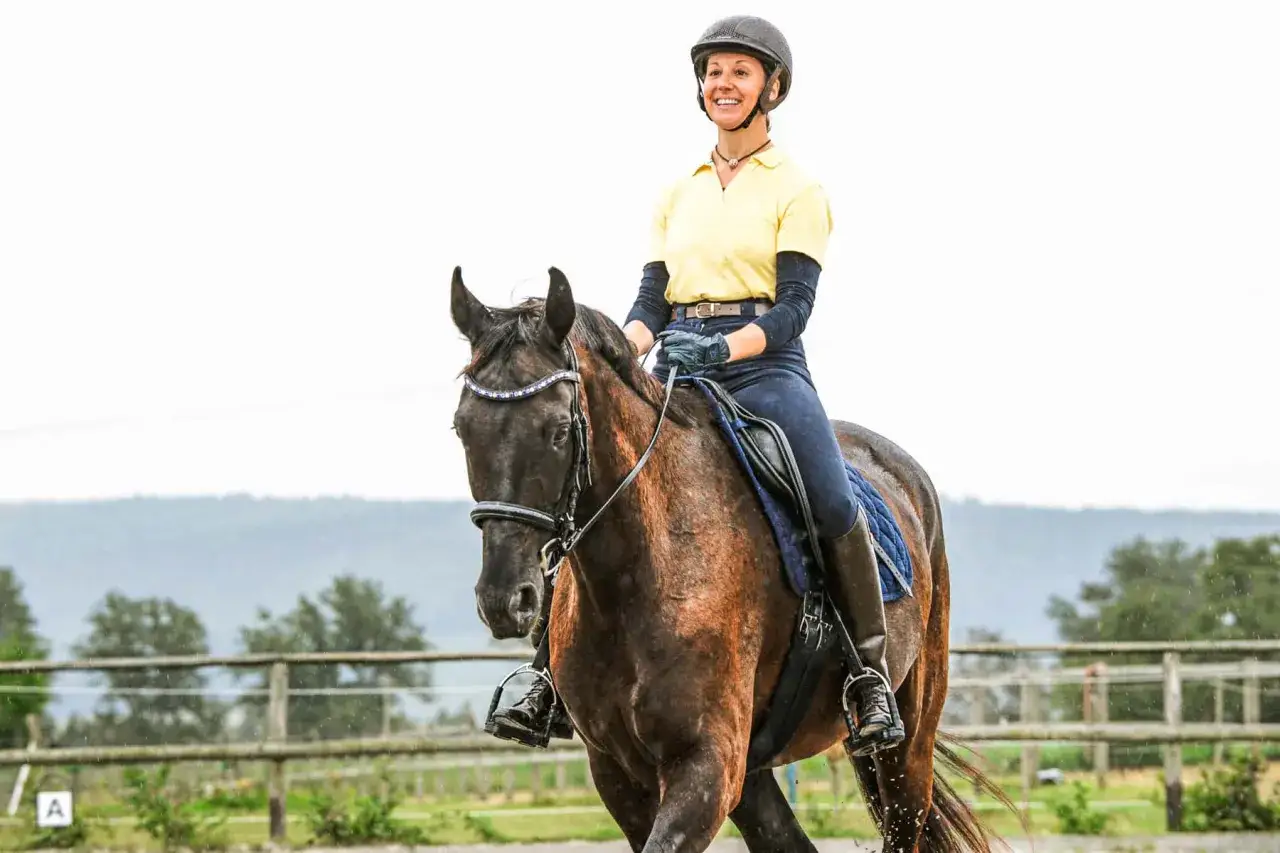 Eine Reiterin in gelbem Shirt und Reithose genießt das Aussitzen beim Reiten auf einem braunen Pferd.