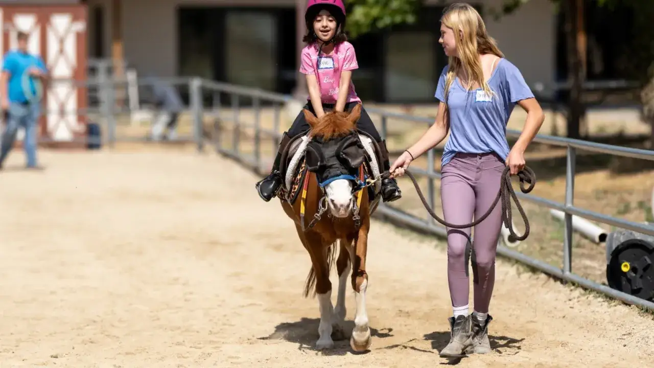 Ein Mädchen lernt reiten, während eine junge Frau das Pony führt. Die Frage, wie lange braucht man um reiten zu lernen, hängt von vielen Faktoren ab.